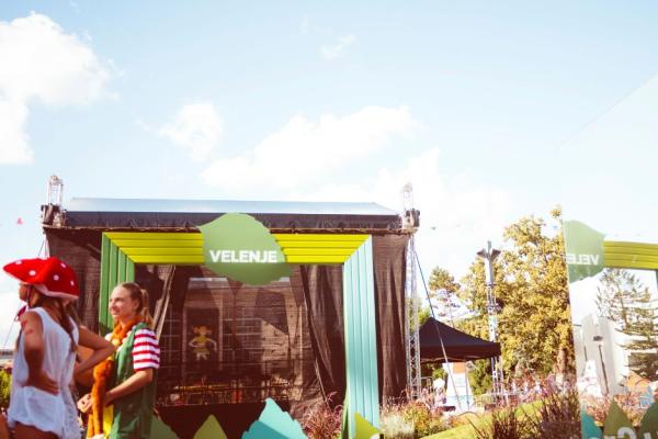 Outdoor event stage with "EU Green Leaf" and "Velenje 2024" banners. Several individuals are in the foreground, one wearing a mushroom costume. Clear blue sky and trees in the background.